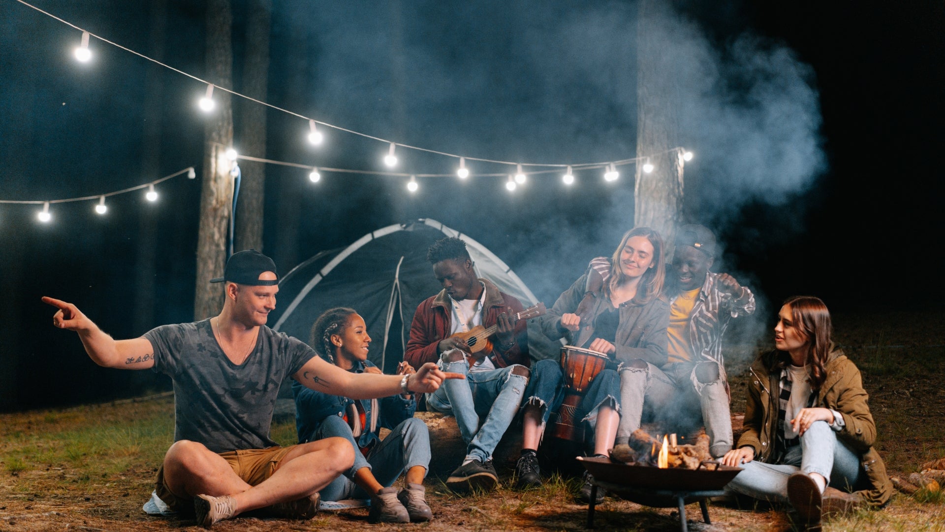 Group of friends enjoying campfire and music under string lights at night